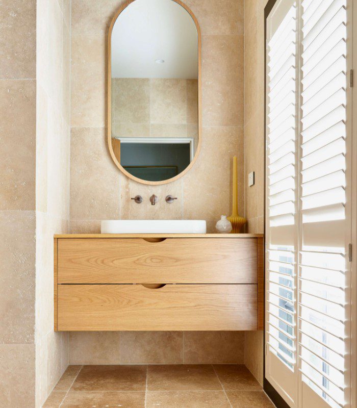 Vistala Travertine is pictured creating a beautiful backdrop for a wooden mirrored bathroom vanity with stainless steel tap features. Large Venetian blinds showcase the limestone’s hues with natural lighting.