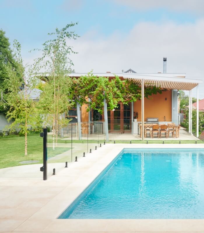 An outdoor pool area featuring a blue natural limestone tiled pool, light cream coloured limestone floors, a lawn with trees, and an orange home in the background with a table and chairs.