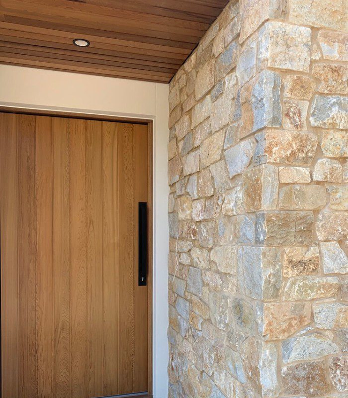 A Peninsula limestone corner wall at the front of a modern home. There’s a timber door with a black handle, and a timber ceiling.