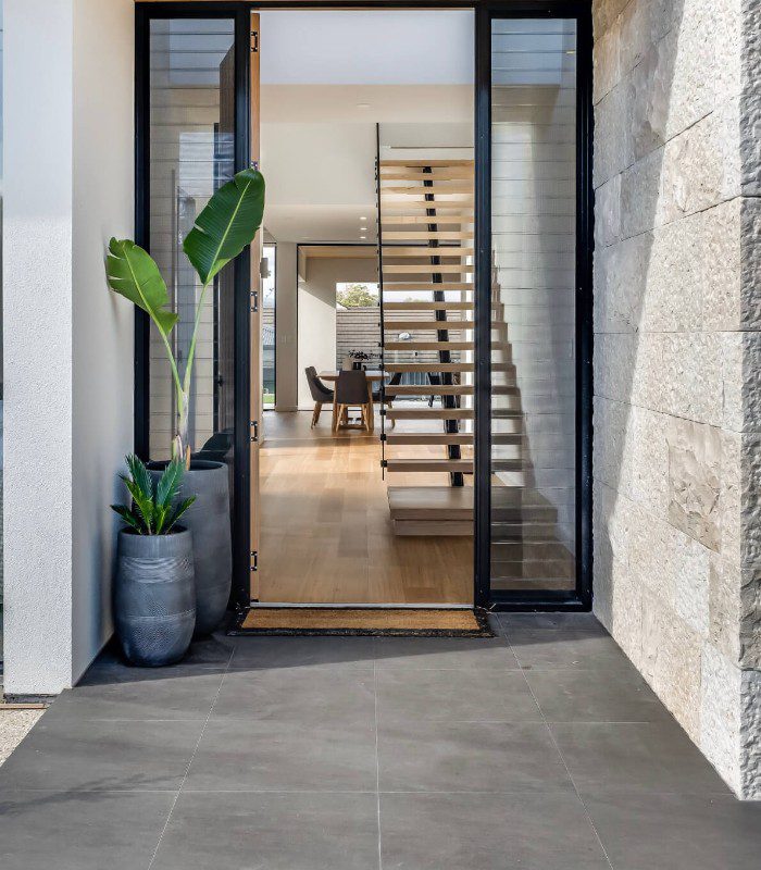 This image shows an entrance to a home shot from the outside through an open door. The outside wall is limestone stone, and the indoor floor covering and staircase are timber. There are pot plants and black door frames to contrast the walling.
