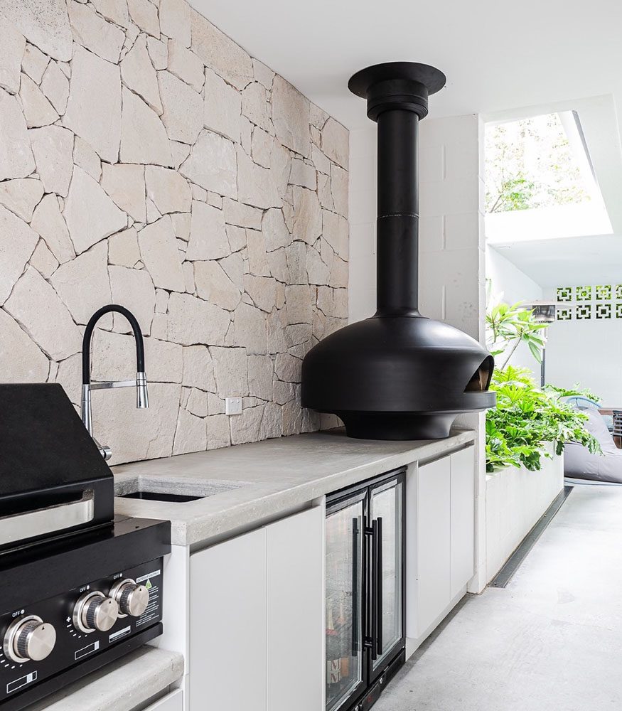 Outdoor kitchen with a concrete bench and white drawers. A black wood oven and pizza oven sit in front of a large dry-stacked Tumbled Beau Sandstone wall. Behind the kitchen area, there is a large lush plant in a white planter box.