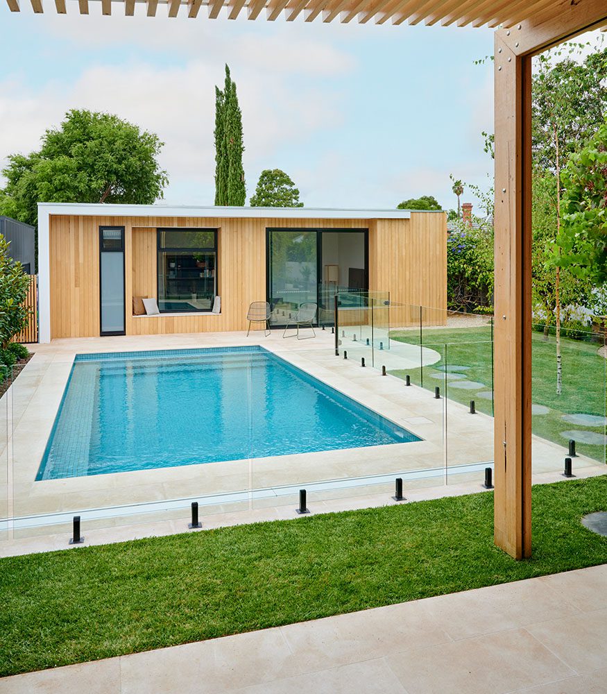 Cream Brulee Limestone tiles around an outdoor pool. A modern wooden home is in the background. The home’s wall indents to form a bench, next to two grey chairs.