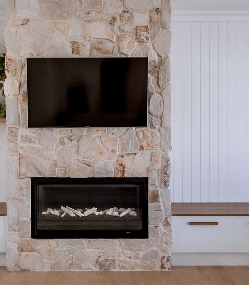 An indoor fireplace featuring white grouted Byron Irregular Sandstone. The stones are rugged, angular and neutral. A TV hangs above the fireplace and sits next to a wooden bench.
