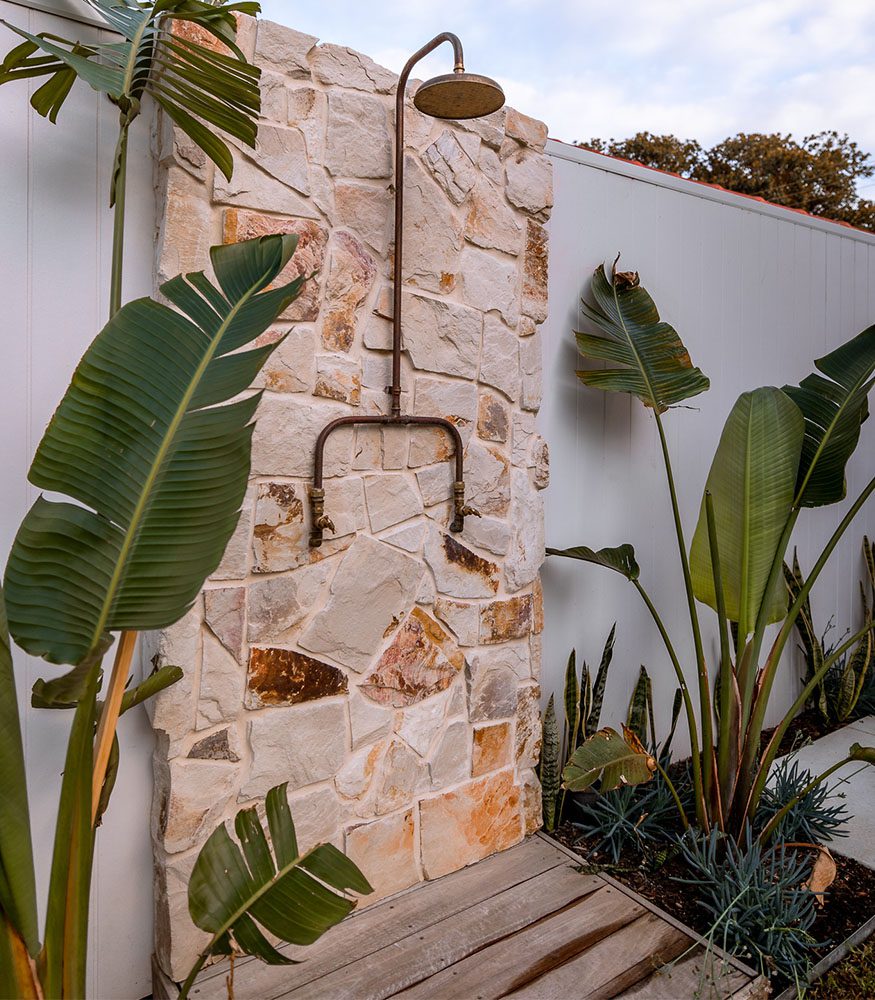 An outdoor shower with a toffee, cream and grey-coloured Byron Irregular Sandstone backdrop. The shower rests against a white wooden slat fence and is surrounded by leafy plants.