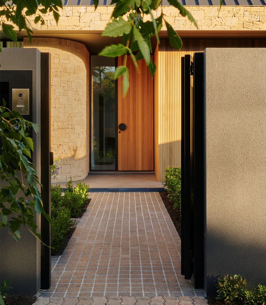 Outdoor granite tiles leading from an open gate set between a home's concrete walls towards a home. The dark Tumbled Nero Cobbles contrast with the wooden front door, and lighter wall tiles.