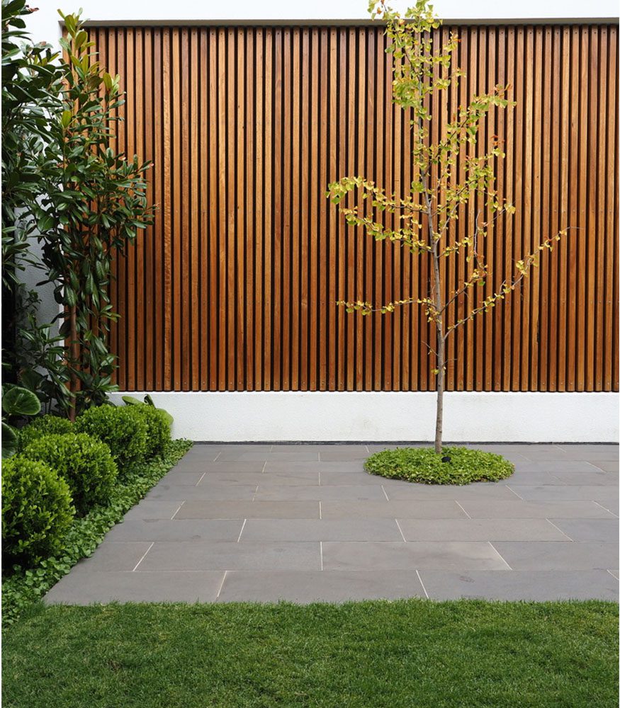 Natural wooden partition behind an Obsidian Bluestone paver patio. Grass borders the pavers and leads to a small bunch of bushes.