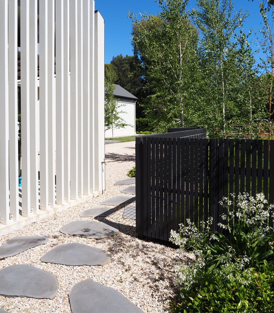 A pathway of smooth Tumbled Obsidian Bluestone steppers leads past a white statement fence and a short black fence. Tall trees surround the backyard.
