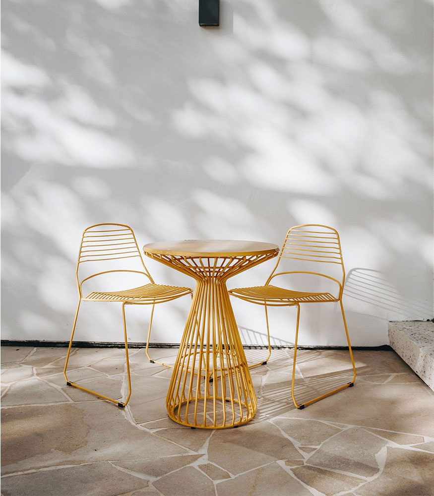 A set of yellow outdoor stools around a high table. The wall is painted white and the floor is caramel limestone in a crazy pattern with white grout.
