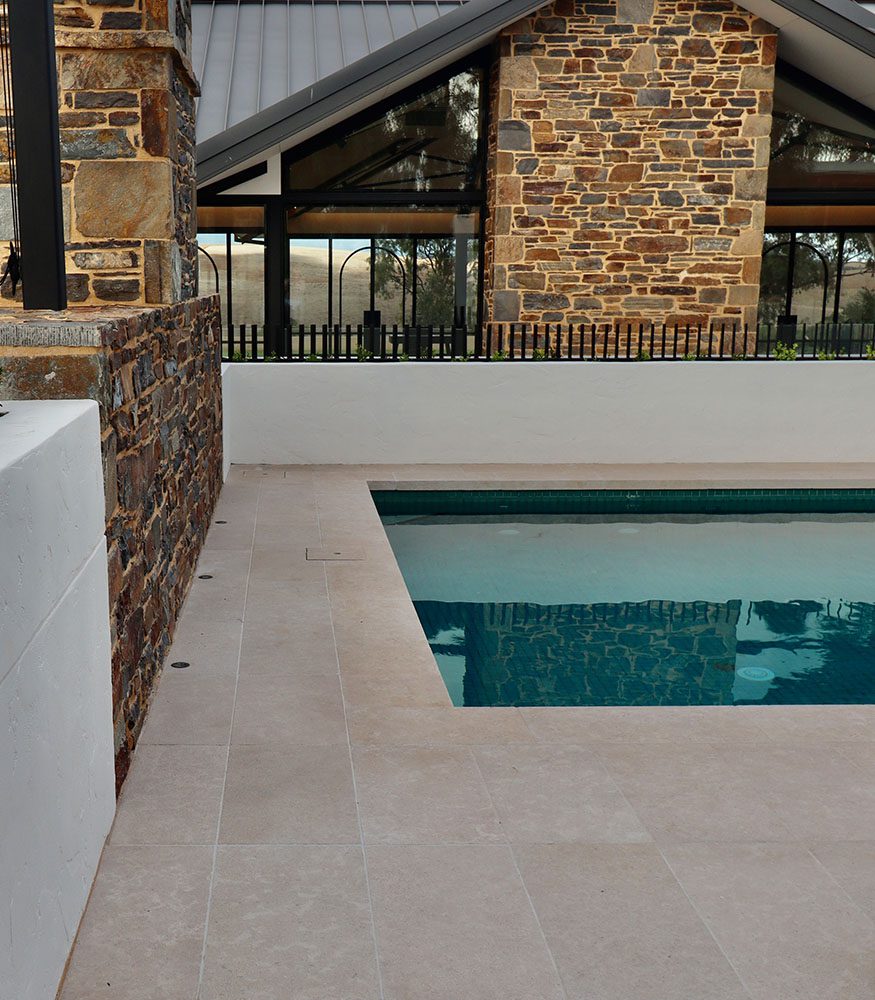 An outdoor pool surrounded with caramel limestone pavers in a grid pattern. Surrounding the pool is a multicoloured stone home with a black roof.