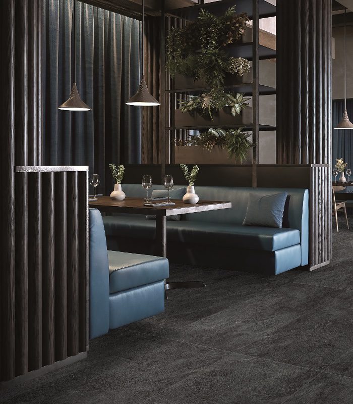 A dark modern restaurant uses bream-coloured Rocky Porcelain floor tiles. Two teal couches face a dark wooden dining table with potted plants on top. Behind one of the couches are shelves with lush hanging plants. Dark wood slat panelling gives the room texture.