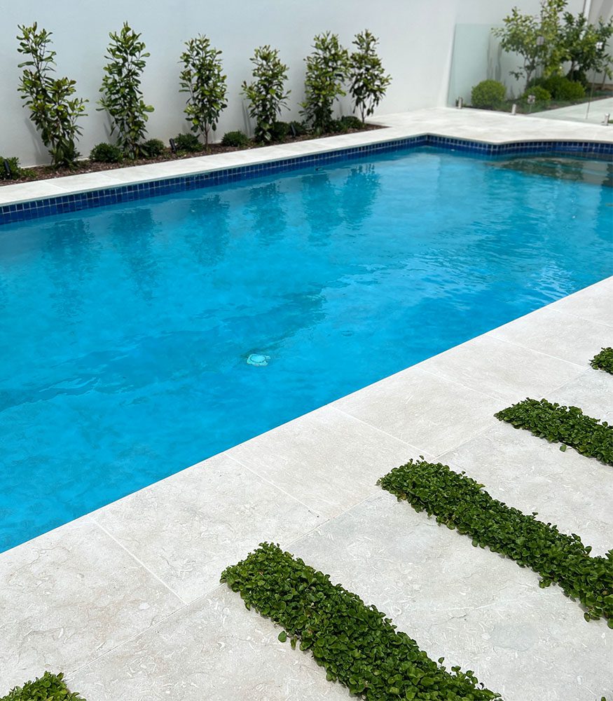 Sage Limestone tiles outline a backyard swimming pool. Shrubbery and glass fencing border the pool.