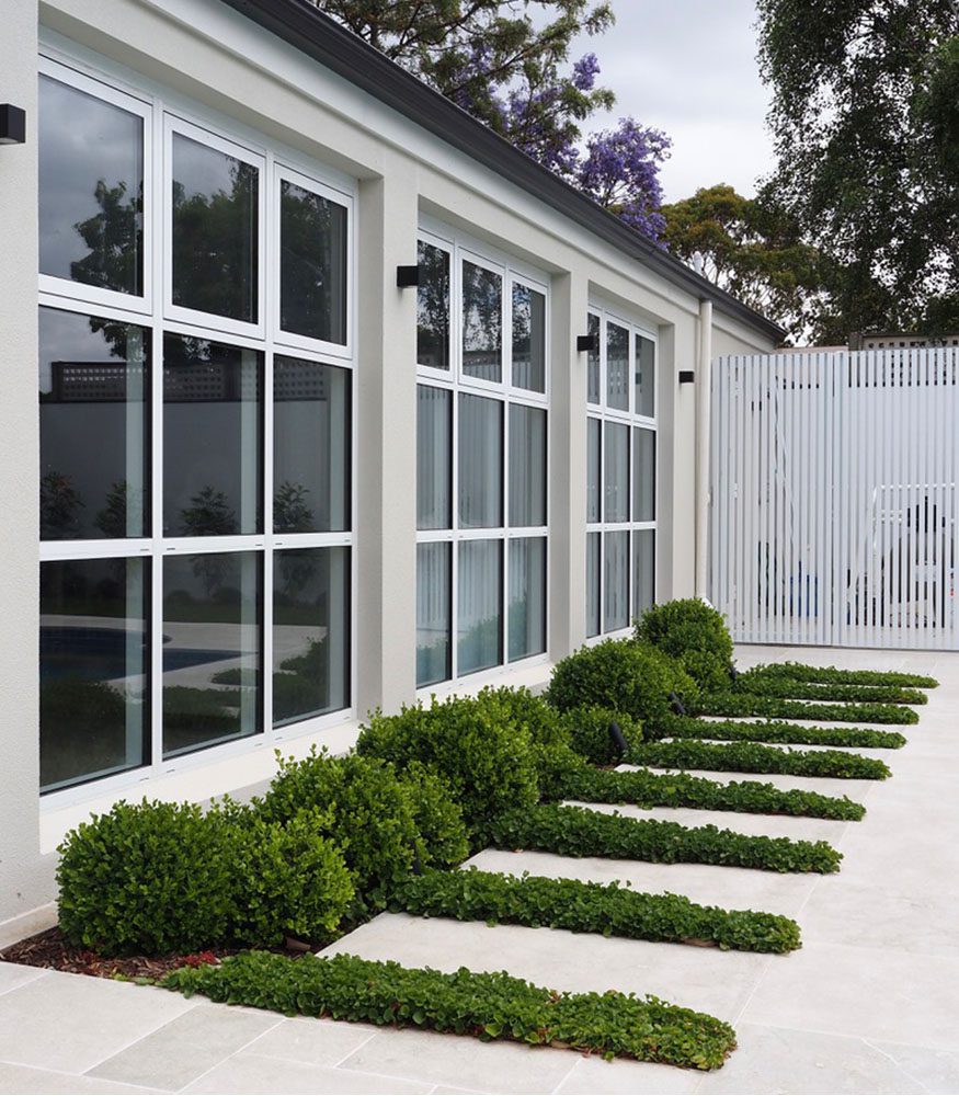 A white-rendered home features large windows in a grid pattern. Short-grown shrubbery is planted along the wall. Tumbled grey Sage Limestone is used to line the ground.