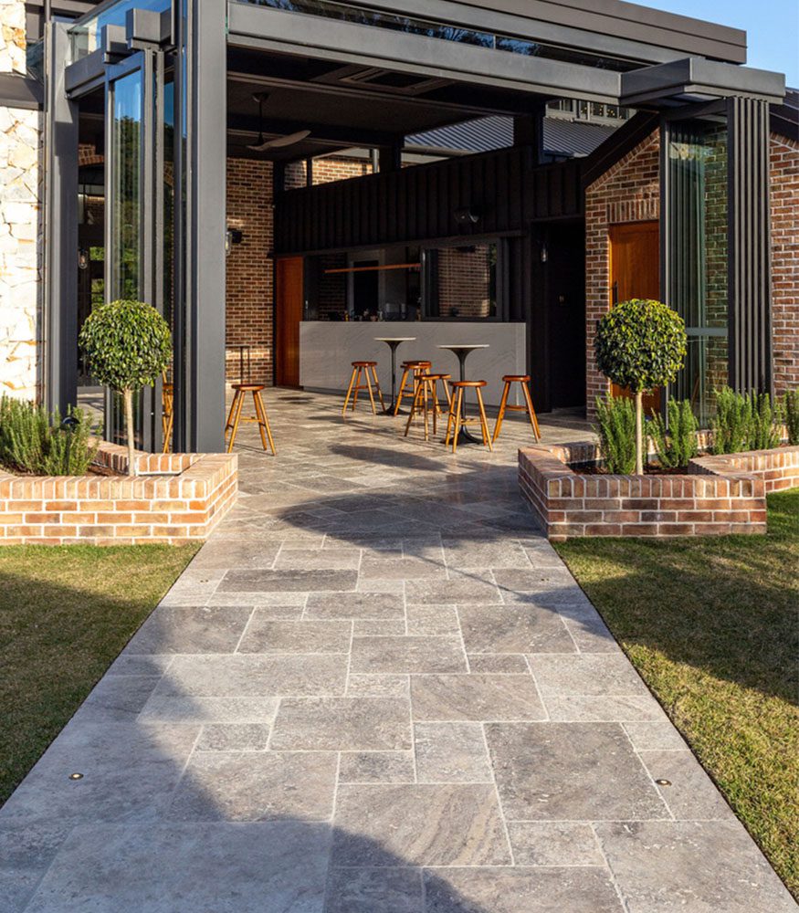 A modern brick building and black veranda. Silver Travertine tiles in a Versailles (French) pattern lead towards and fill the outdoor area. The tiles exhibit grey, brown, and cream hues.