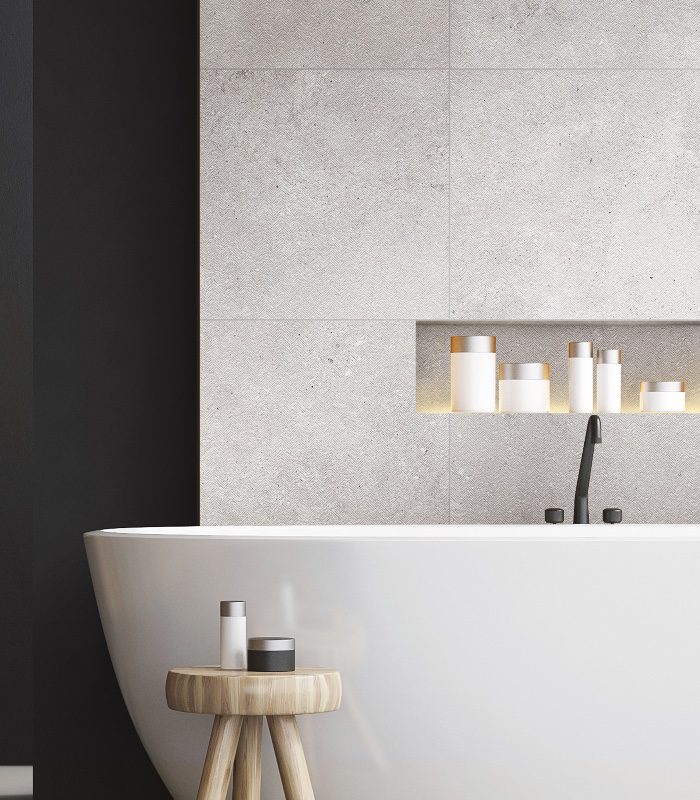 Nuage Porcelain tiles as walling in a bathroom. There’s a freestanding white bathtub in front of the wall, with a black tap, and a small wooden stool holding bath products.