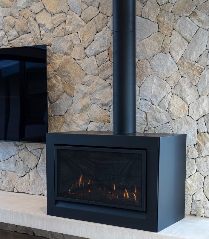 A black freestanding wood-burning fireplace sits to the right of a mounted flatscreen TV. The wall is built with Irregular Somerton Quartz Limestone in a dry stack.